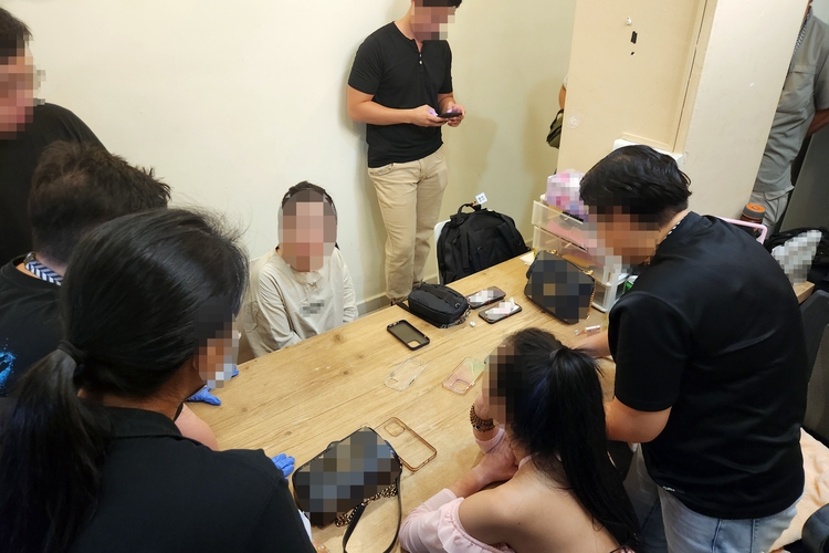 officers surrounding two female suspects seated at a beige wooden dining table, in a well lit room