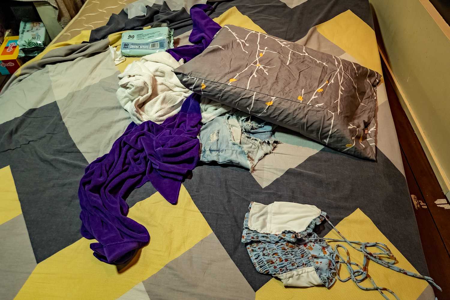 an image of a bed lying on the floor, with articles of clothing left in a messy state and baby wipes boxes beside it