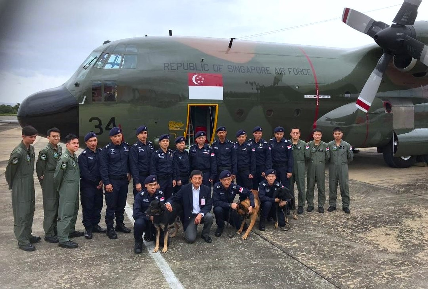 officers standing near the entrance of the C130, on the runway, with someone the ambassador in a blue suit kneeling infront of the group posing for the camera. 