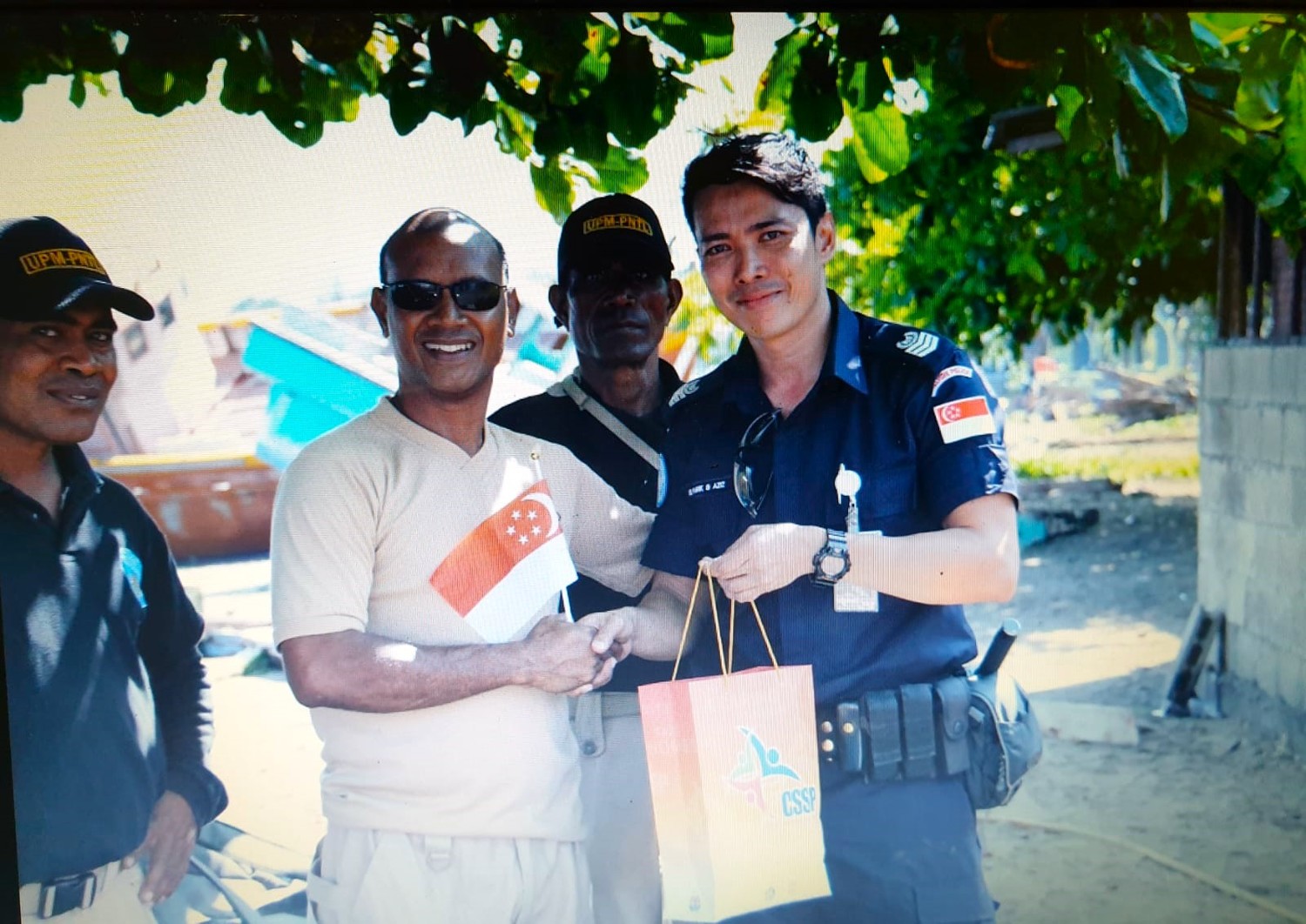 SI Farik on the right, giving a small gift bag to the commissioner, under a shaded tree. The Commissioner is a bald headed man, wearing a pair of sun glasses and a light brown t-shirt. 