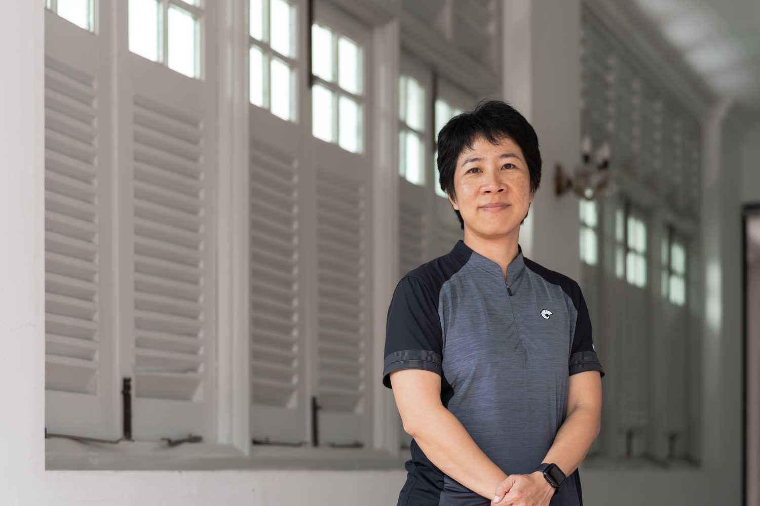 Supt Toh standing in a passage way, with short black hair, grey civilian short sleeve top, with arms infront of her where her left hand is covering her right hand. 