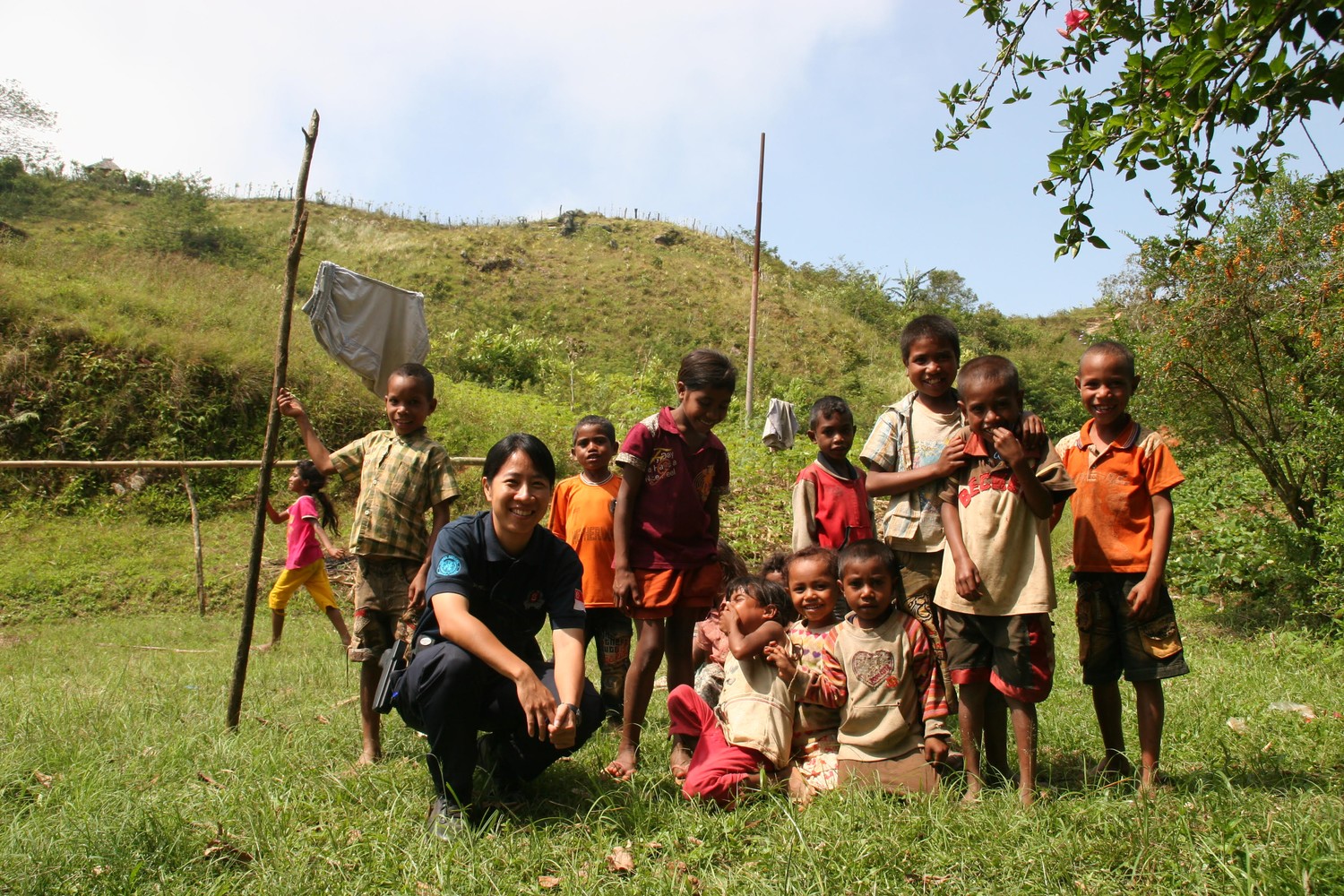 Supt Toh in a crouched position, posing for a camera on a green field, with a few children beside her. On the right is a tree providing shade and in the background is a big green mountain
