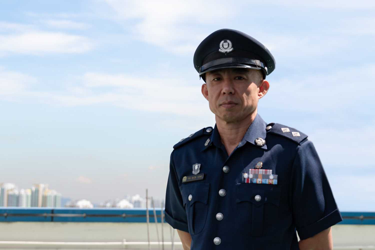 Inspector Tan Buck Song posing for the camera, wearing his Singapore Police Force uniform 3 and peak cap, against the blue sky and white clouds background. 