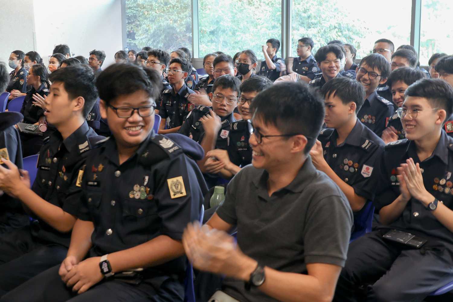 Crowd of NPCC Cadets seated and waiting for award ceremony, a boy is smiling, engaging in conversation with his Teacher Officer. 