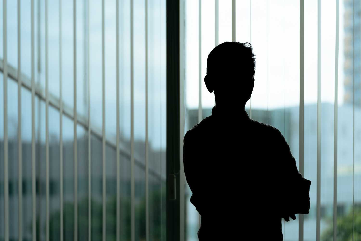 a silhouette of an male figure standing infront of an office window, with the light shining from the back against the window blinds