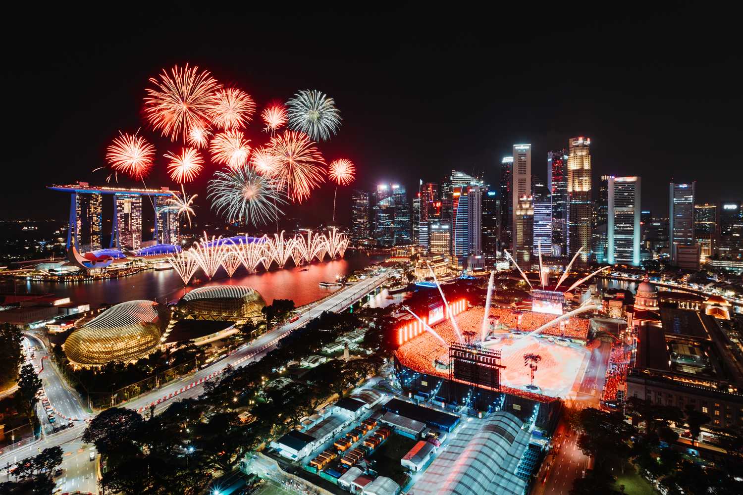 Vibrant red and white fireworks are seen with the skyline of Singapore's iconic buildings like the Marina Bay Sands Hotel in the background. The night sky is filled with the red, blue, white, purple, turquoise, and yellow lights from the buildings surrounding the float.