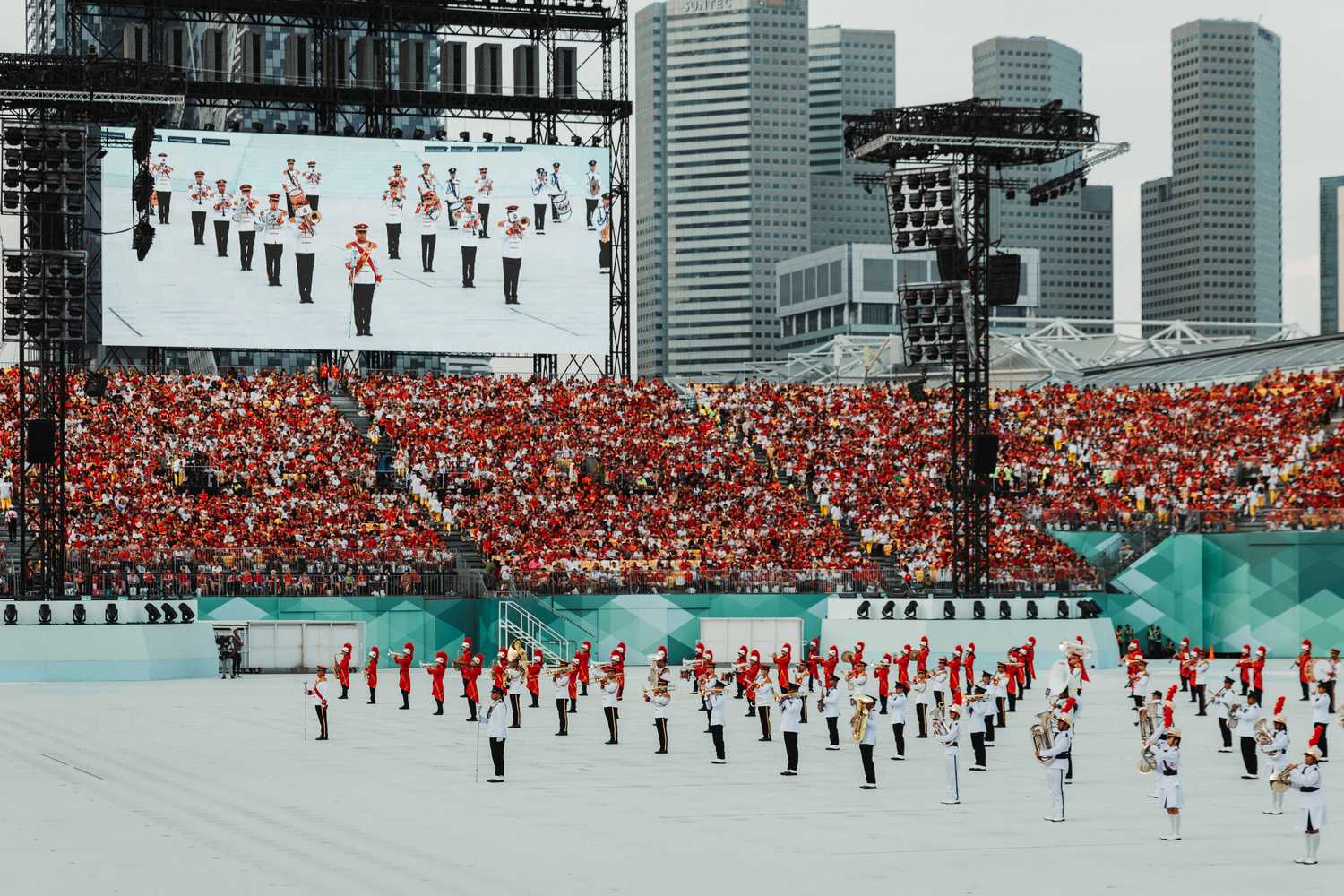The SPF, SAF, and secondary school students from Bowen and Yuhua Secondary School putting up the military band. The stage is white. Some performers are wearing red top and black while others are wearing white uniform with black and white pants. The performers are holding instruments such as the tuba and trumpet. They are standing in alignment.