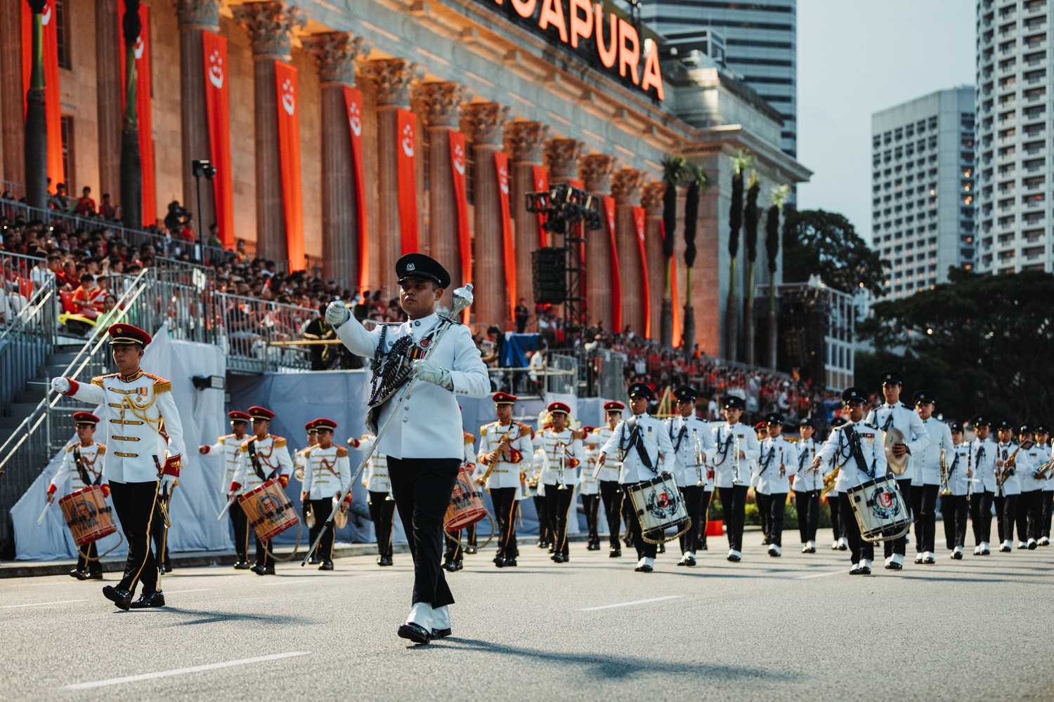 The SPF band marching along the road facing the national gallery of singapore. The officers face's look focused, one man leading his band members who march behind him while playing various instruments such as the bass drum, which has the SPF logo on it.