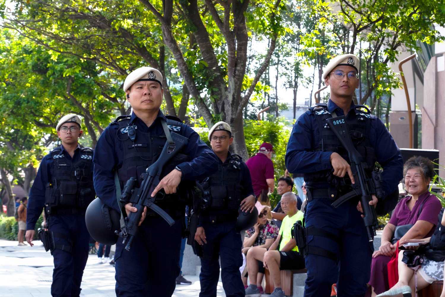 The Protective Security Command (ProCom) In-Situ Reaction Team (IRT) officers patrolling the area surrounding the NDP to ensure everyone's safety. The officers look focused and alert, and are holding on to their Heckler and Koch MP5 submachine gun. There is an elderly lady to the right of the photo seated on a bench smiling as she looks at the officers.