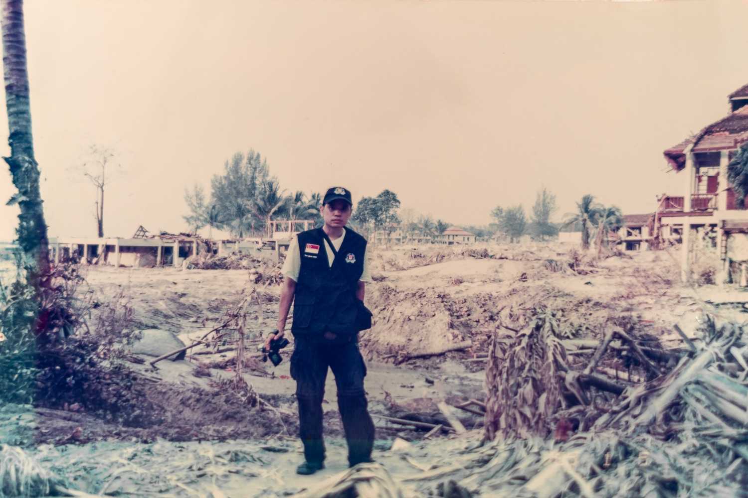 photo of officer wearing blue vest, holding camera in the hand. Behind him is a destroyed landscape with a lot of broken debris, caused by a tsunami