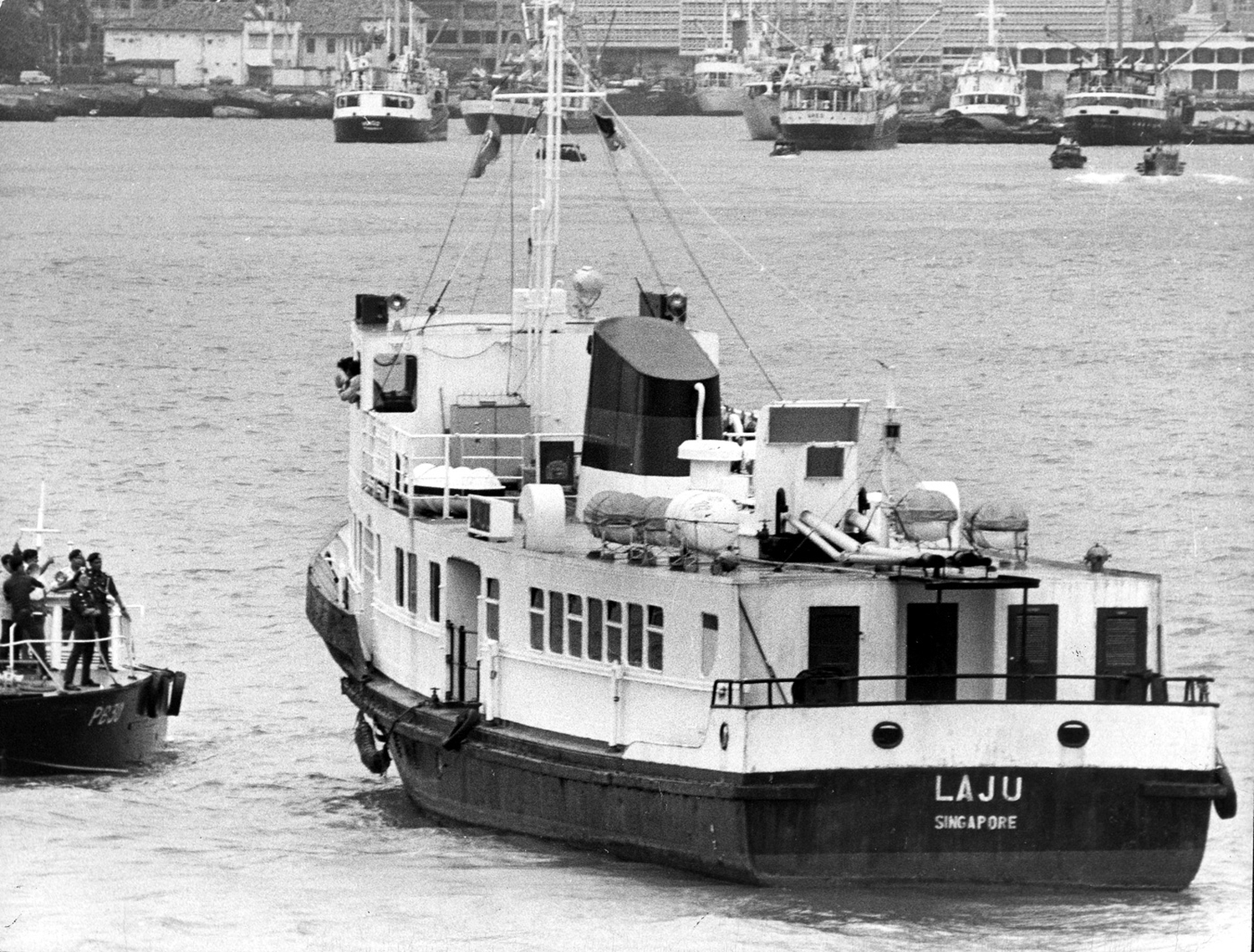 a black and white image of a boat at sea, with a smaller boat beside. The smaller boat has a police officer talking towards the big boat, Laju. 