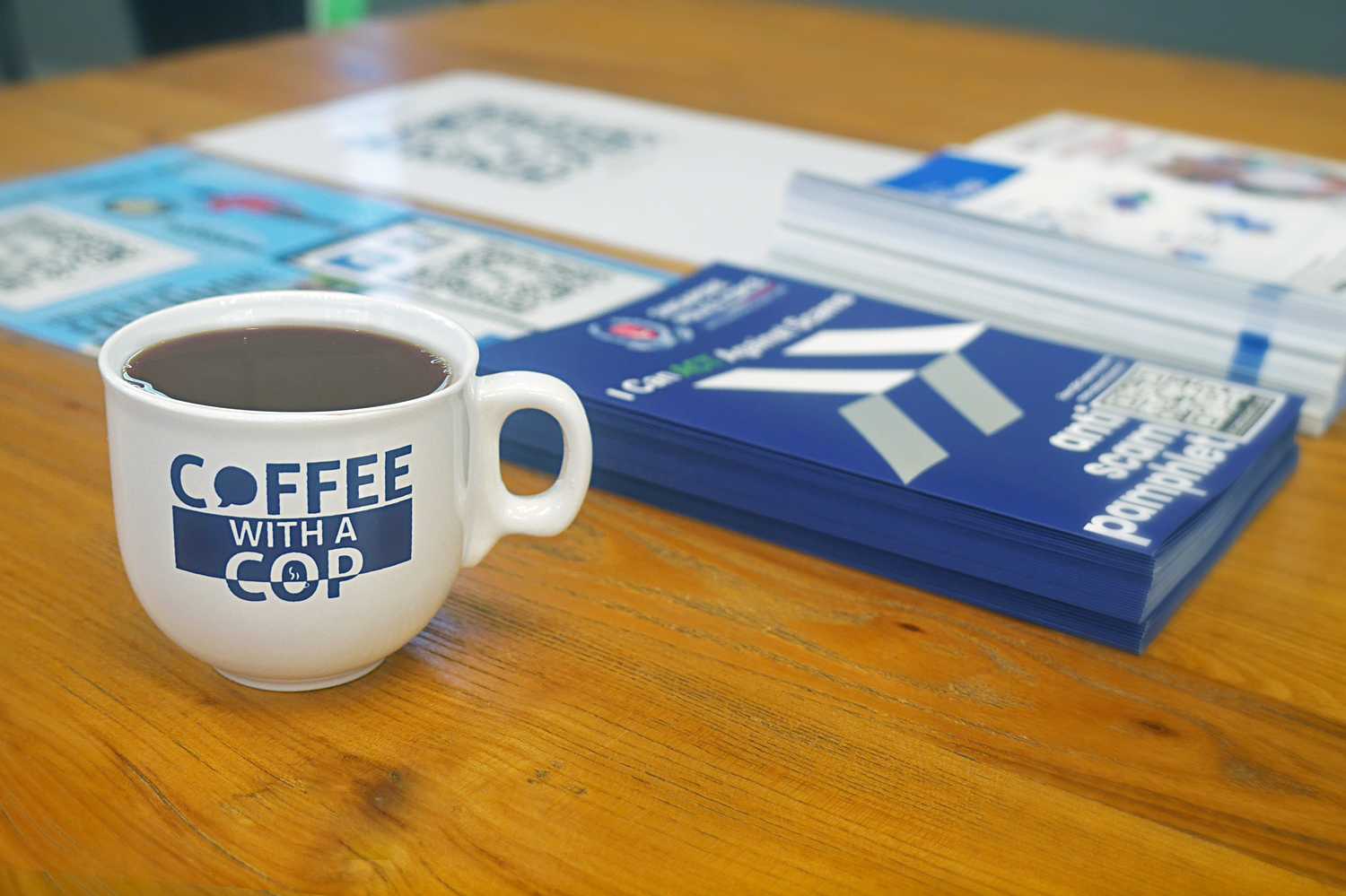 A coffee with a cop cup is placed on a wooden table beside a Police Anti-Scam pamphlet.