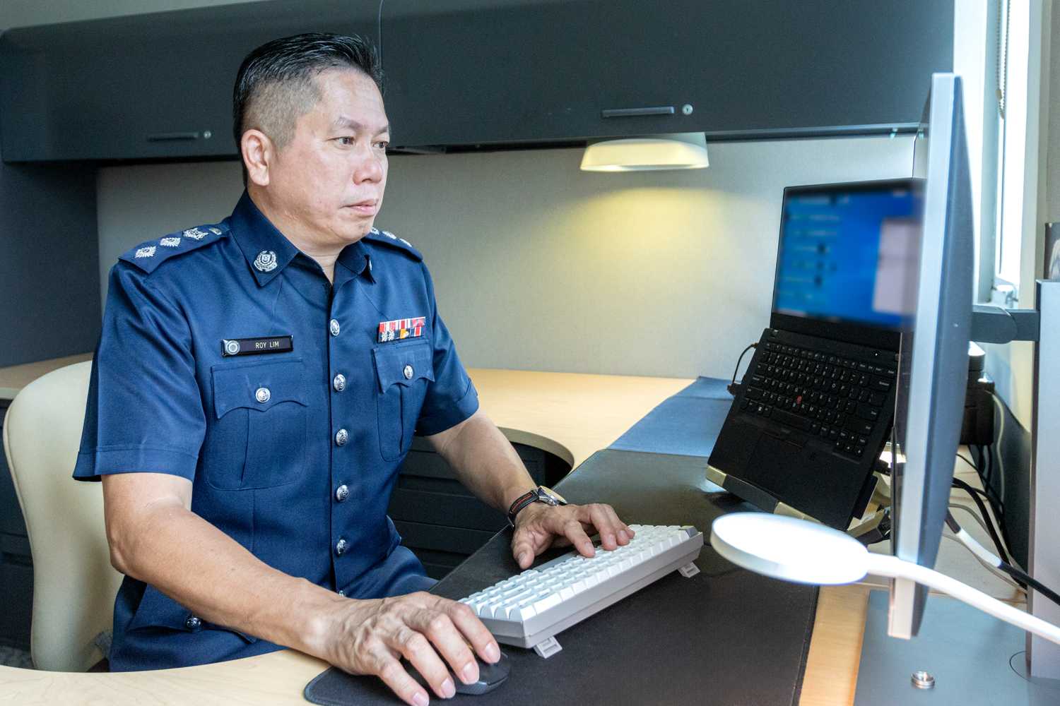 Superintendent of Police (Supt) Roy Lim is sitting on a desk and typing away on a keyboard with his left hand. His right hand holds the mouse. He is looking intently at the computer screen.