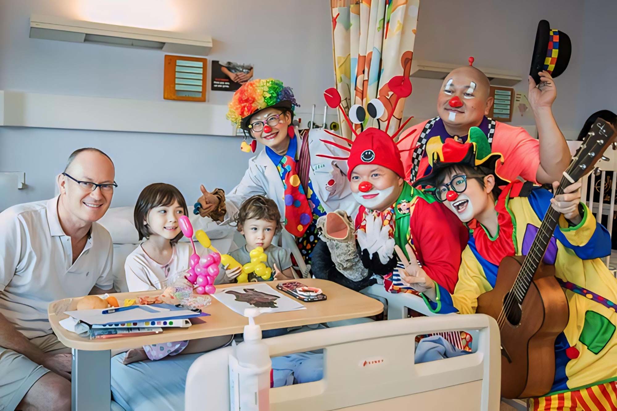 a group of clowns standing and cheerfully entertaining two hospital patients who is with their parent