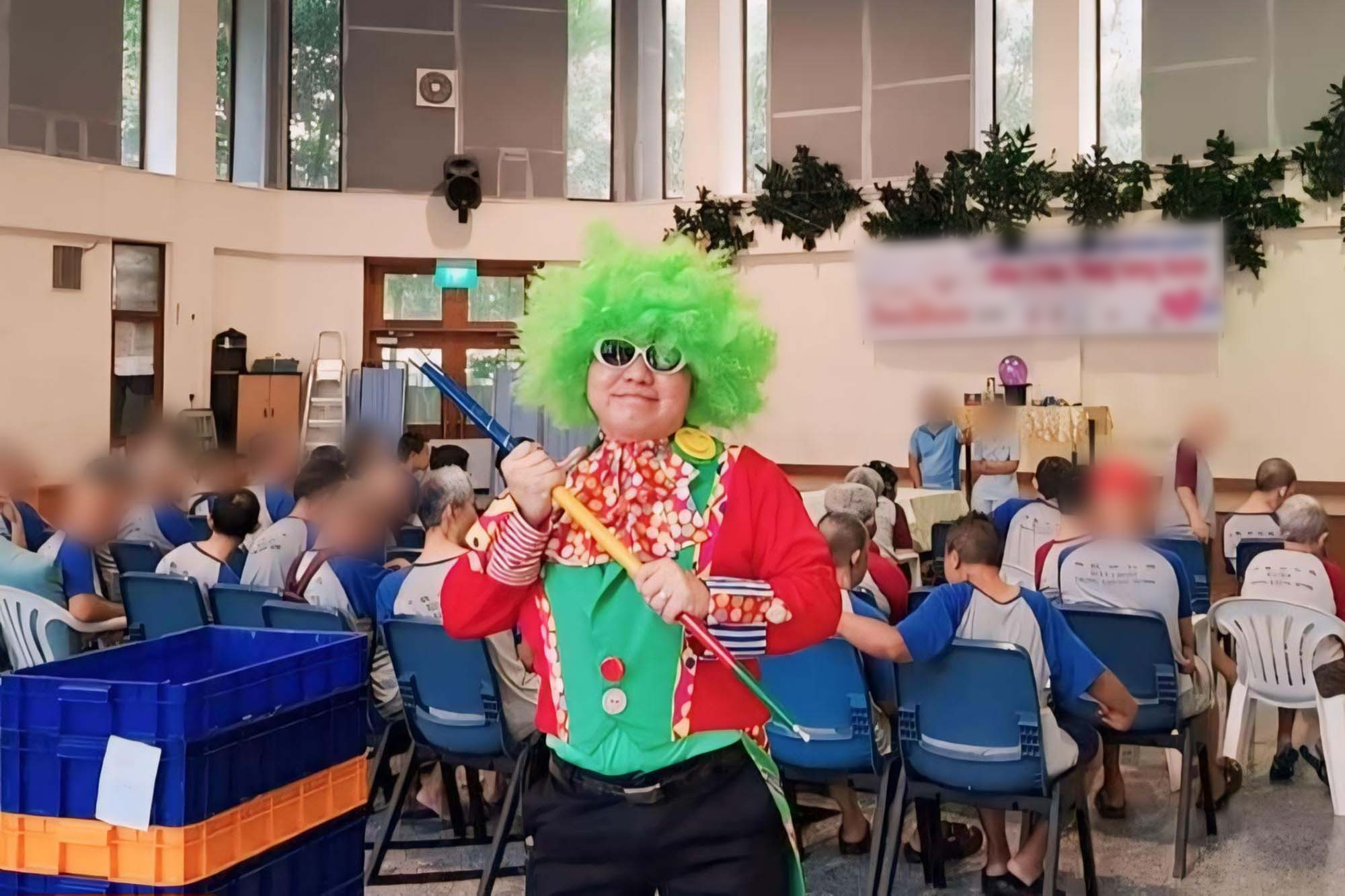 DSP jeremy ang dressed with a colourful wig standing and posing for the camera, with a lot of old people behind him in a nursing home setting