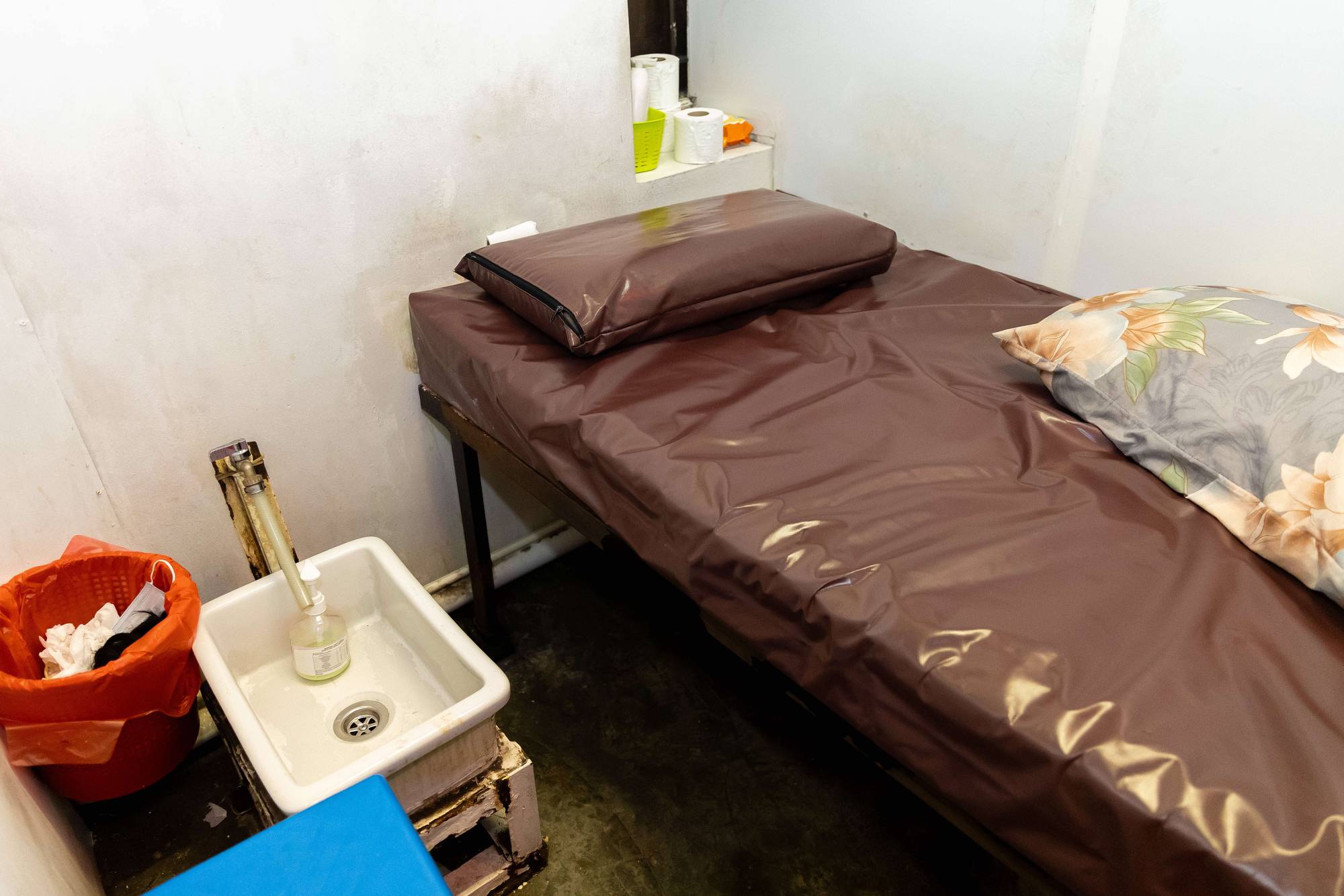 the room contains a small high bed with PVC covering on a brown bed. Beside there's a small basin that is at ankle height, and a small red waste bin