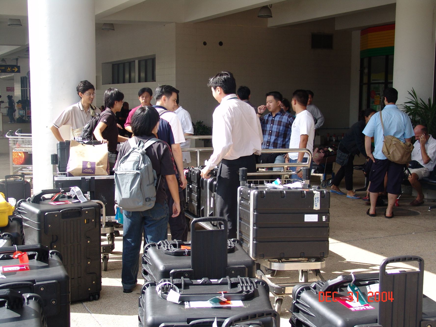 a shot of the officers at the entrance of the phuket airport with a lot of luggage around them. The scene looks messy and cluttered